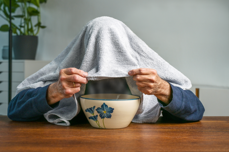 Eine Person mit einem Handtuch über dem Kopf hält eine Schüssel mit Blumenmuster auf einem Tisch.