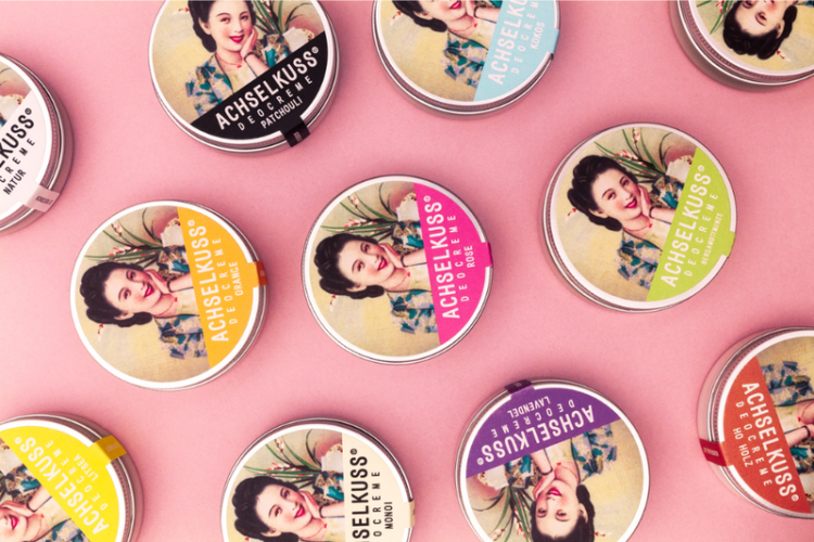 Bunte Dosen mit verschiedenen Düften der Marke ACHSELKUSS auf einem rosa Hintergrund angeordnet.