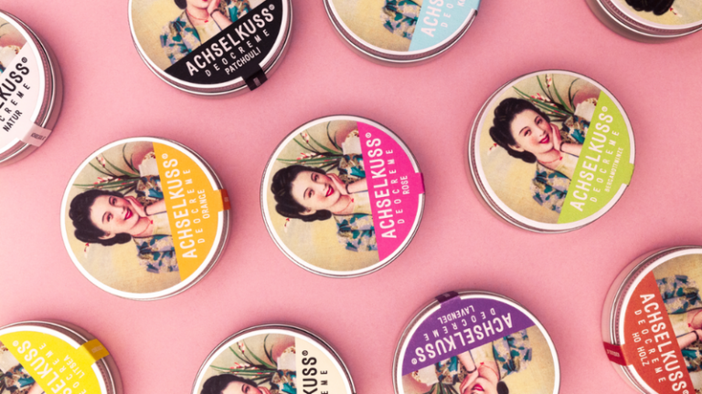 Bunte Dosen mit verschiedenen Düften der Marke ACHSELKUSS auf einem rosa Hintergrund angeordnet.