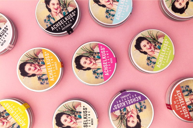 Achselkuss.png Bunte Dosen mit verschiedenen Düften der Marke ACHSELKUSS auf einem rosa Hintergrund angeordnet.