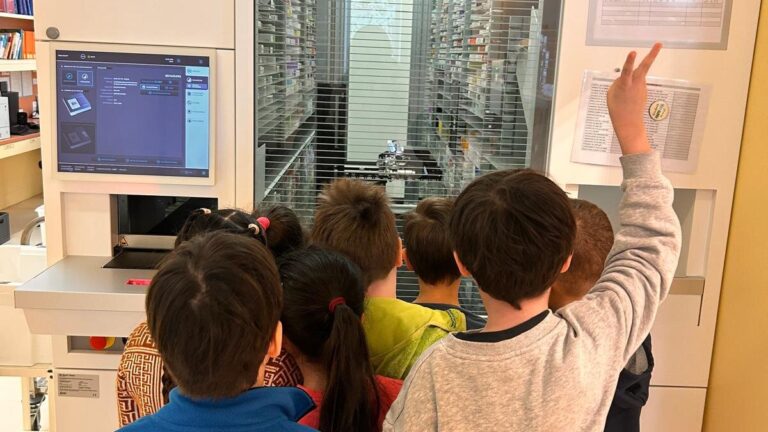 Gruppe von Kindern steht vor einem automatisierten Medikamentenschrank mit Bildschirm und Informationen.