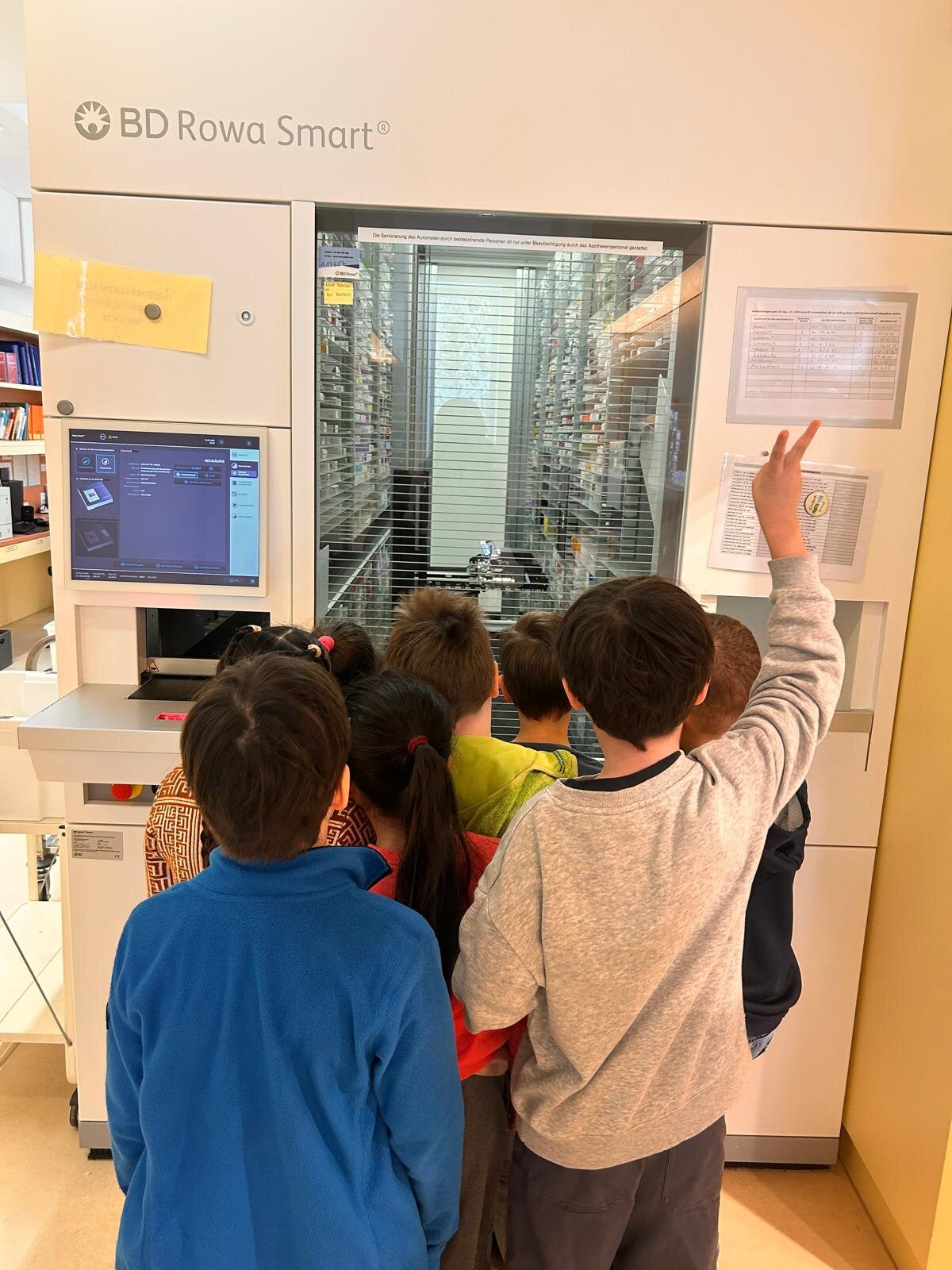 vorschule-hadersdorf-besuch.jpeg Gruppe von Kindern steht vor einem automatisierten Medikamentenschrank mit Bildschirm und Informationen.