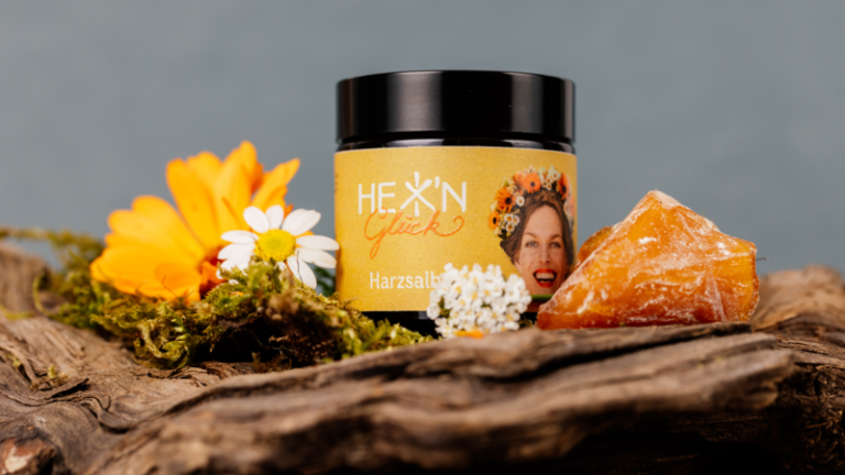 Ein Glas mit Gelben Harzsalz auf einem Holzteller, umgeben von bunten Blumen und einer Harzstück.