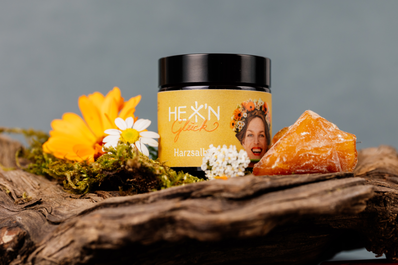 Ein Glas mit Gelben Harzsalz auf einem Holzteller, umgeben von bunten Blumen und einer Harzstück.