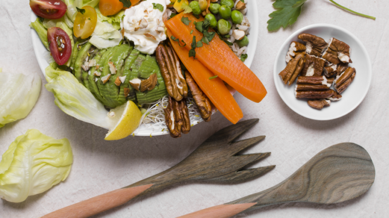 Bunte Salatschüssel mit Avocado, Karotten, Erbsen, Mais, Tomaten und Nüssen, begleitet von Holzbesteck und einer Schale Nüssen.