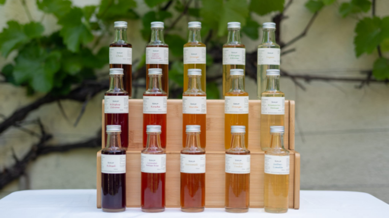 Eine Anordnung von Glasflaschen in verschiedenen Farben auf einem Holzregal, beschriftet mit Namen von Essenzen.