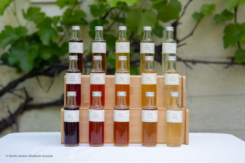 Eine Anordnung von Glasflaschen in verschiedenen Farben auf einem Holzregal, beschriftet mit Namen von Essenzen.