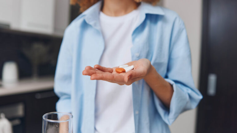 Eine Person hält zwei Kapseln in einer Hand, während in der anderen ein Glas Wasser steht, in einer modernen Küche.